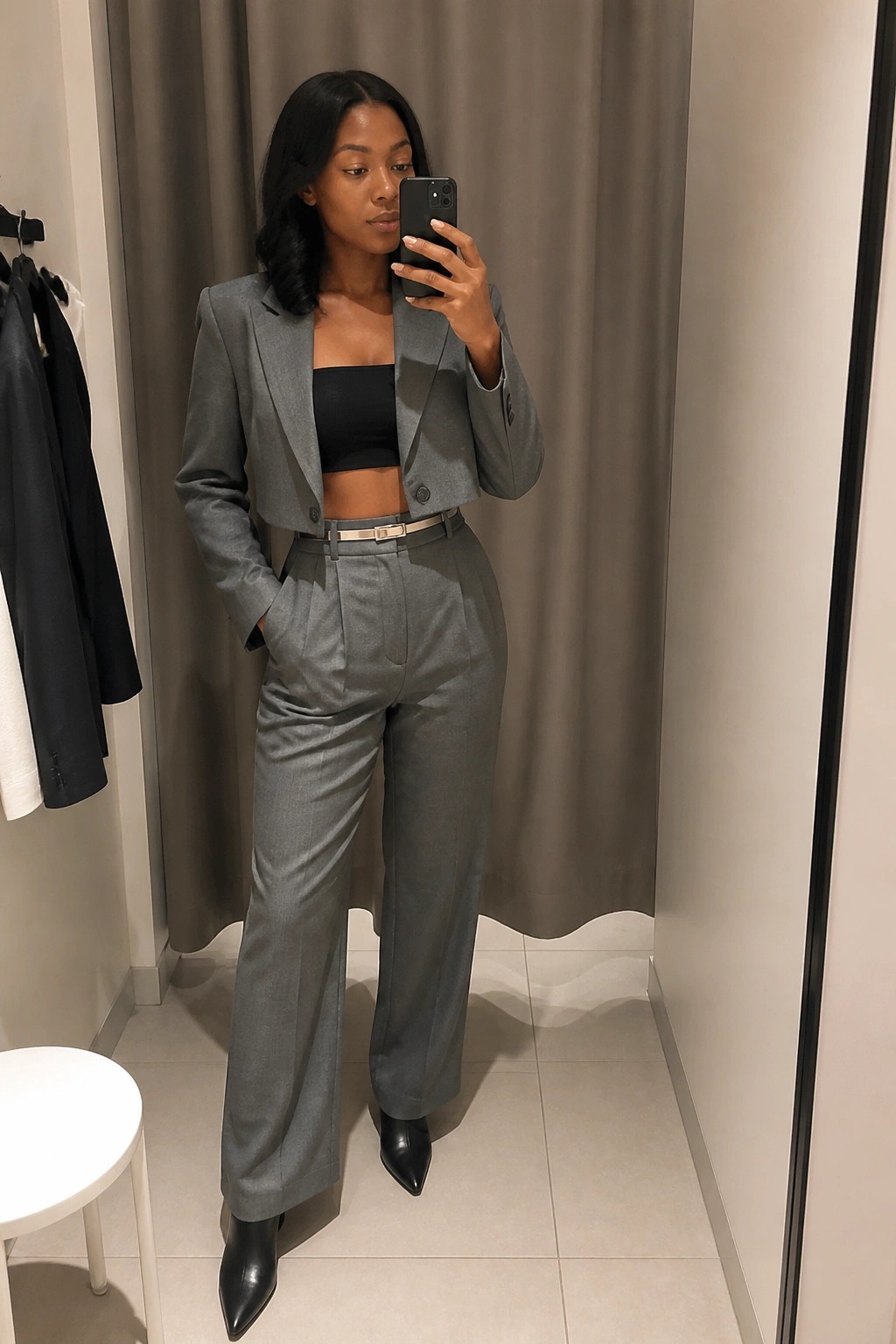 Woman in gray cropped blazer and trousers taking mirror selfie in fitting room