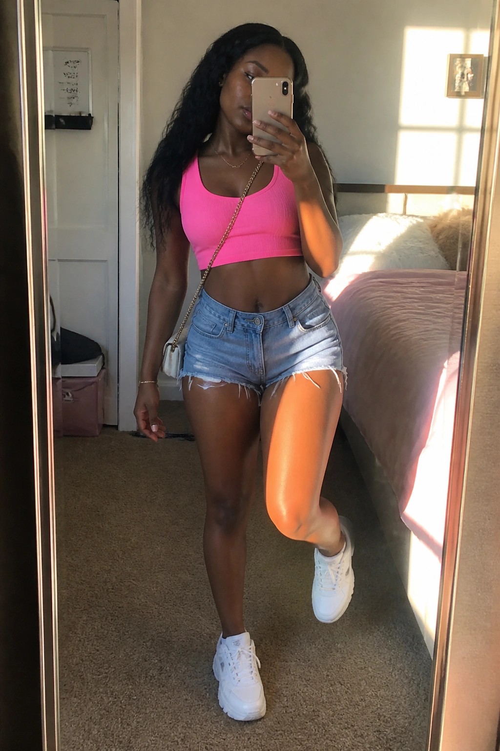 Woman in bright pink ribbed crop top, light blue frayed denim shorts, white chunky sneakers, and gold chain crossbody bag posing in mirror selfie.