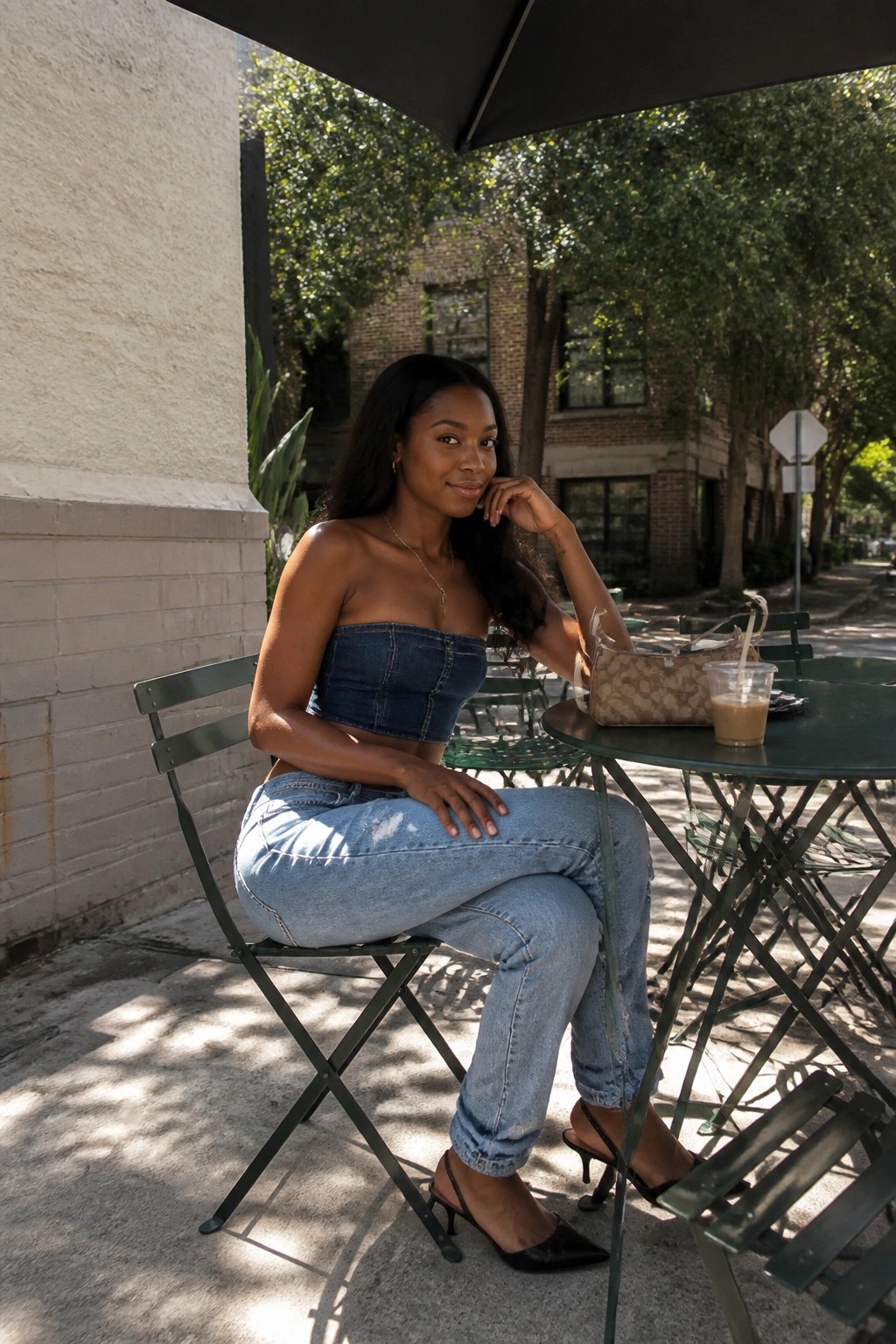 Woman in denim crop top and jeans at green outdoor cafe table with drink