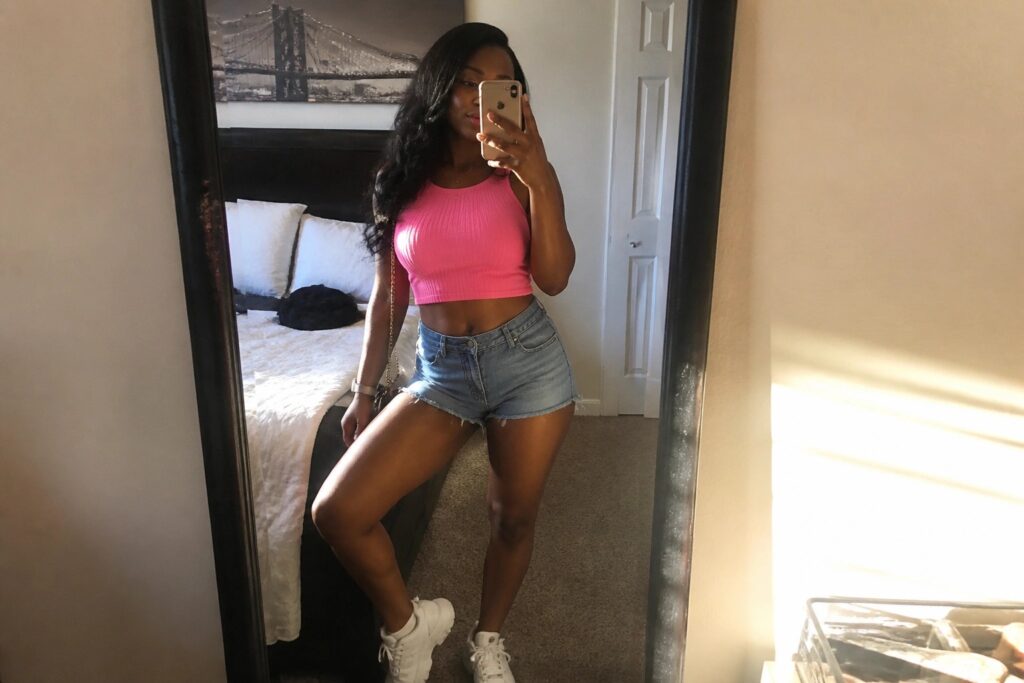 20 Baddie Miami Outfits for Black Women With Bold Street Style Energy Woman in bright pink ribbed crop top, light blue frayed denim shorts, white chunky sneakers, and gold chain crossbody bag posing in mirror selfie.