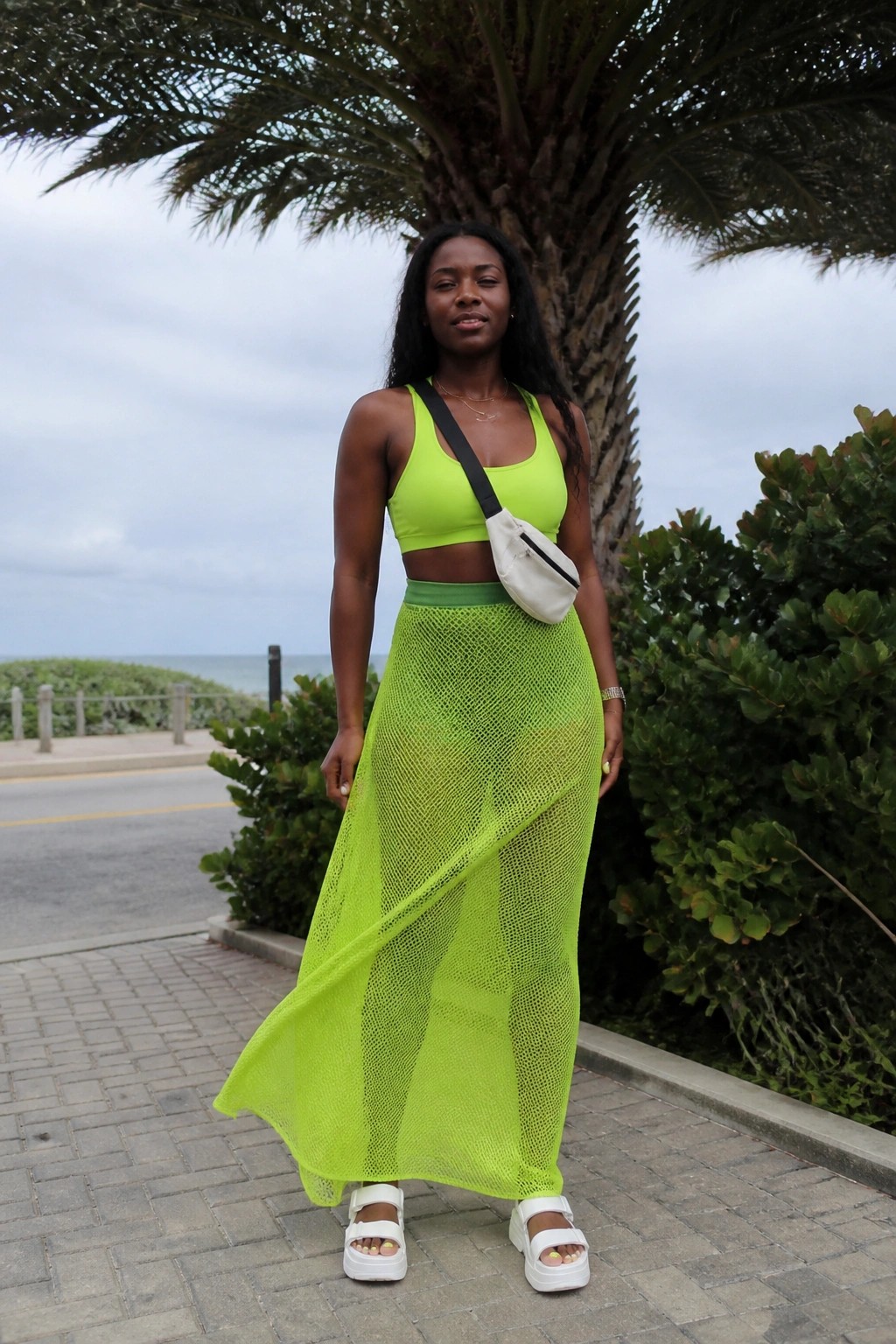 Woman in neon green crop top and sheer mesh skirt beside palm tree by ocean