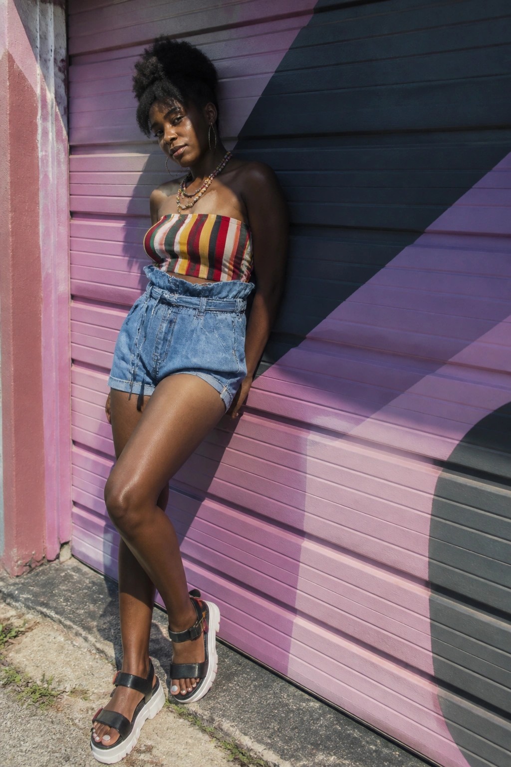 Woman in striped crop top and denim shorts leaning on colorful shutter door