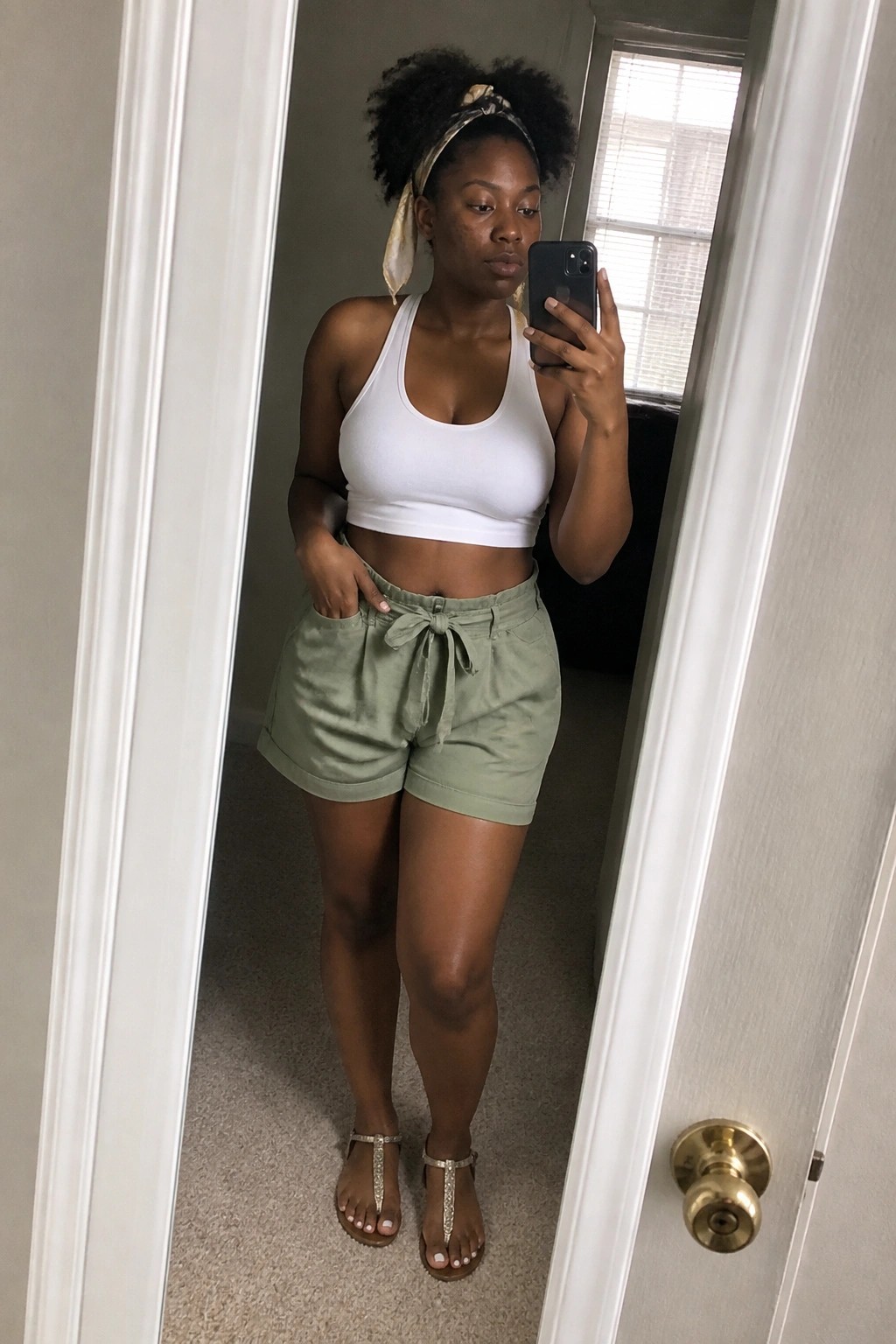 Woman wearing white ribbed crop tank top with sage green high waisted tie front shorts and embellished sandals taking mirror selfie in doorway