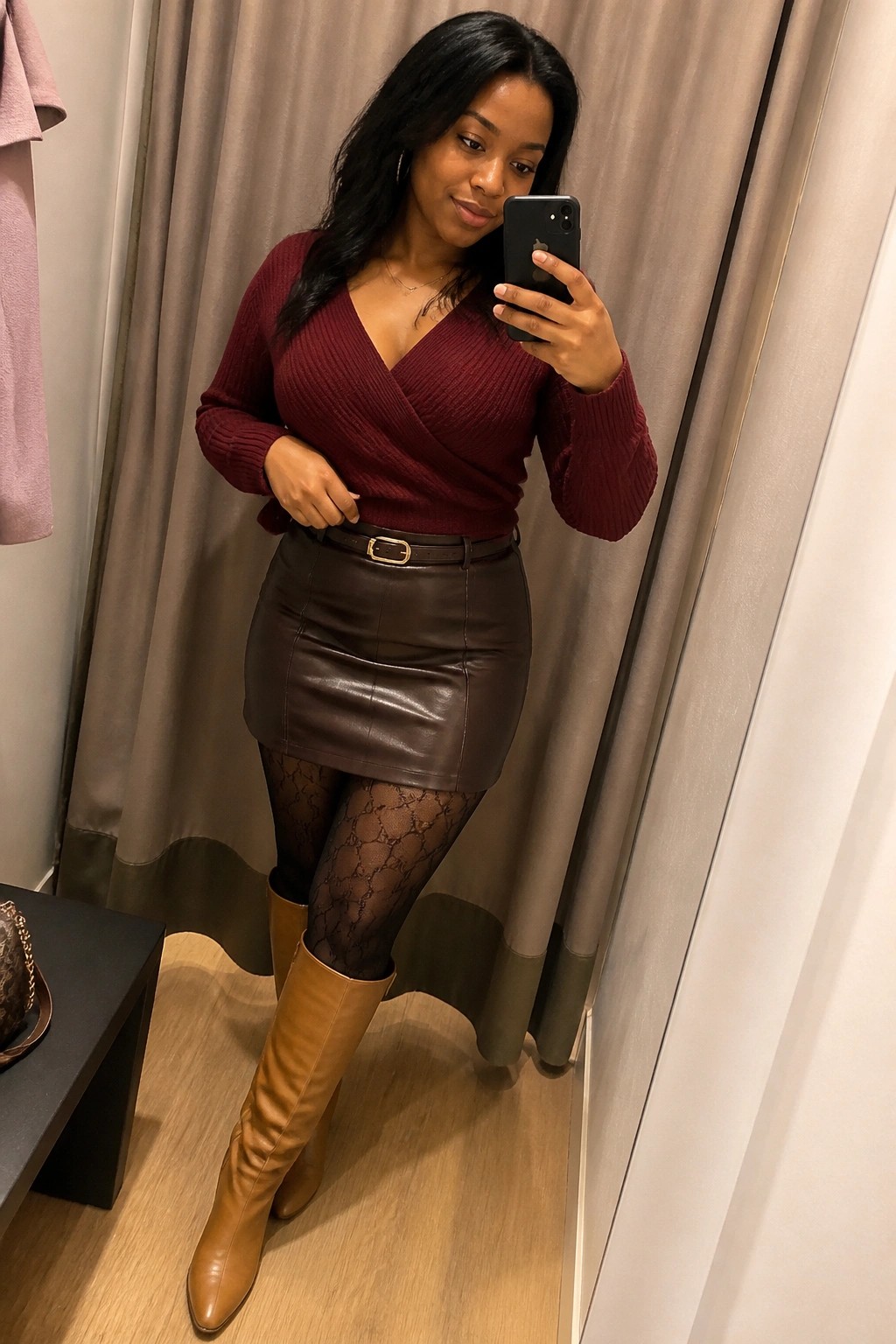 Woman in maroon wrap sweater and brown leather skirt taking mirror selfie in fitting room.