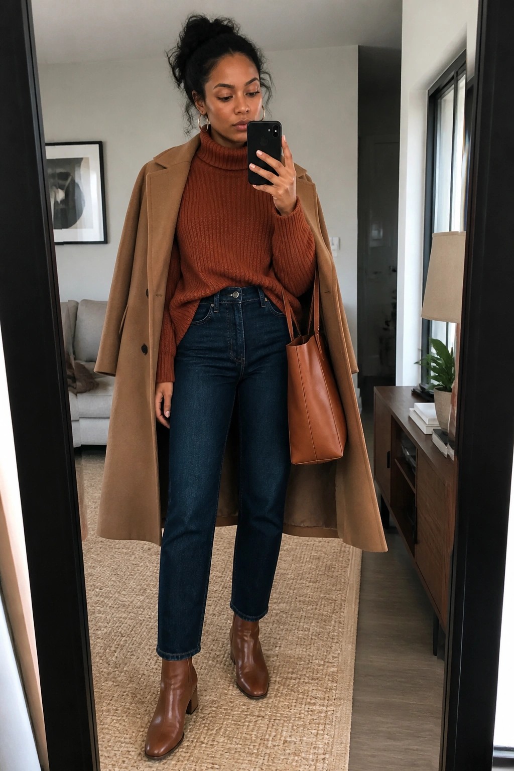 Woman in camel coat over rust turtleneck taking mirror selfie with brown tote