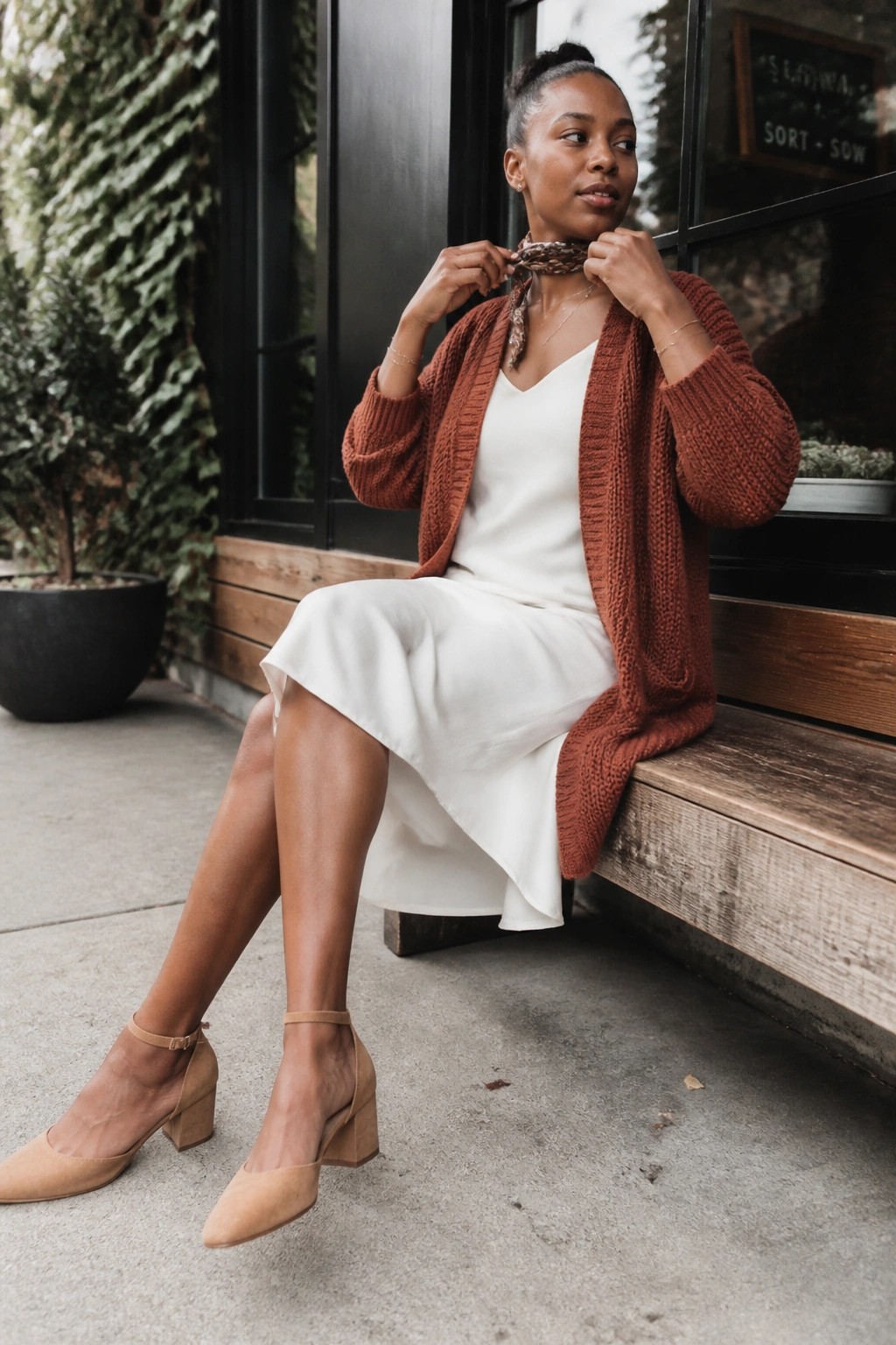 A Black woman seated outdoors wearing a rust orange chunky knit cardigan over a white V-neck midi dress paired with a patterned neck scarf, beige block heel shoes, and delicate gold jewelry.