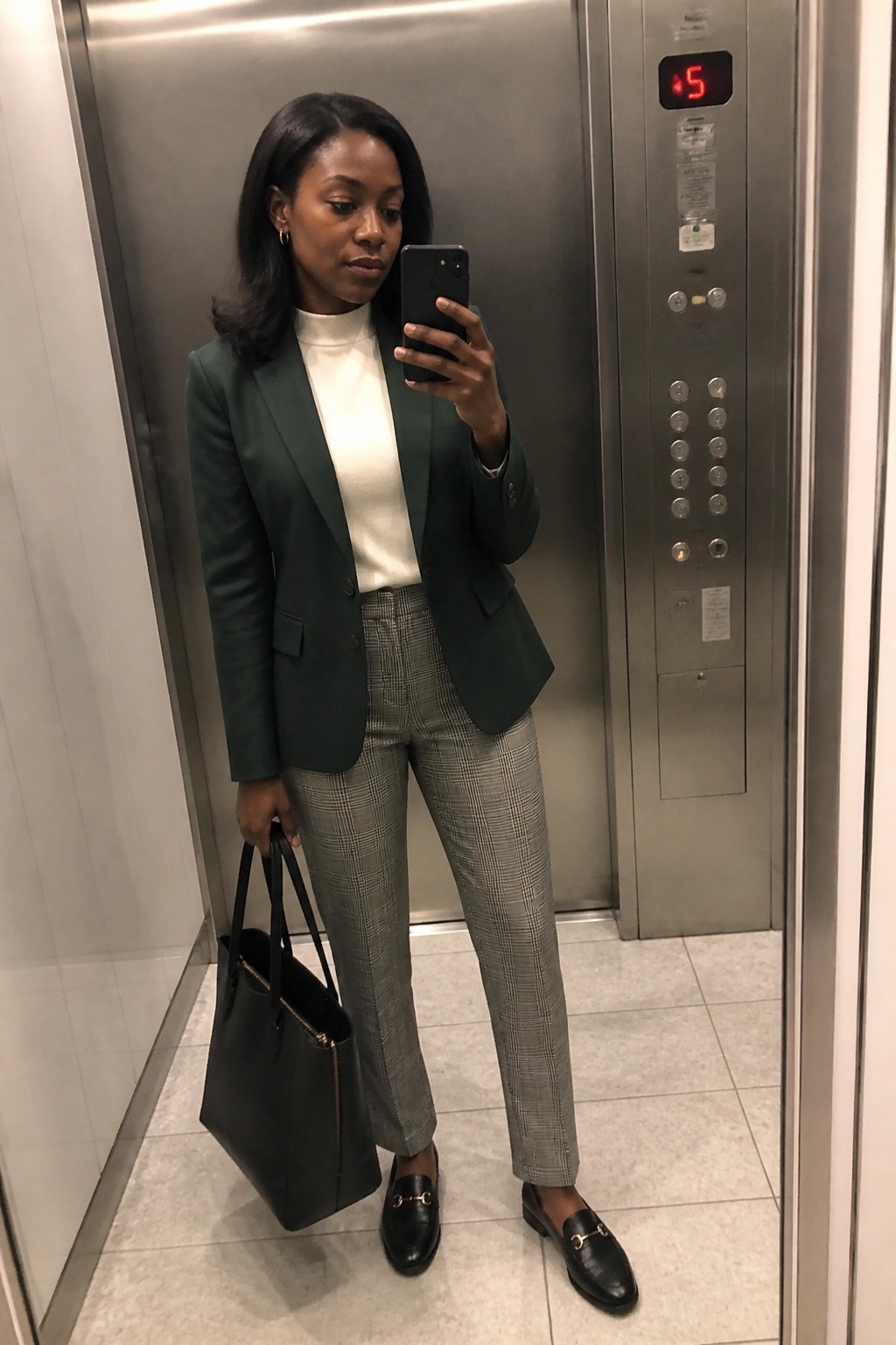 Woman in green blazer and plaid pants taking selfie in elevator with bag