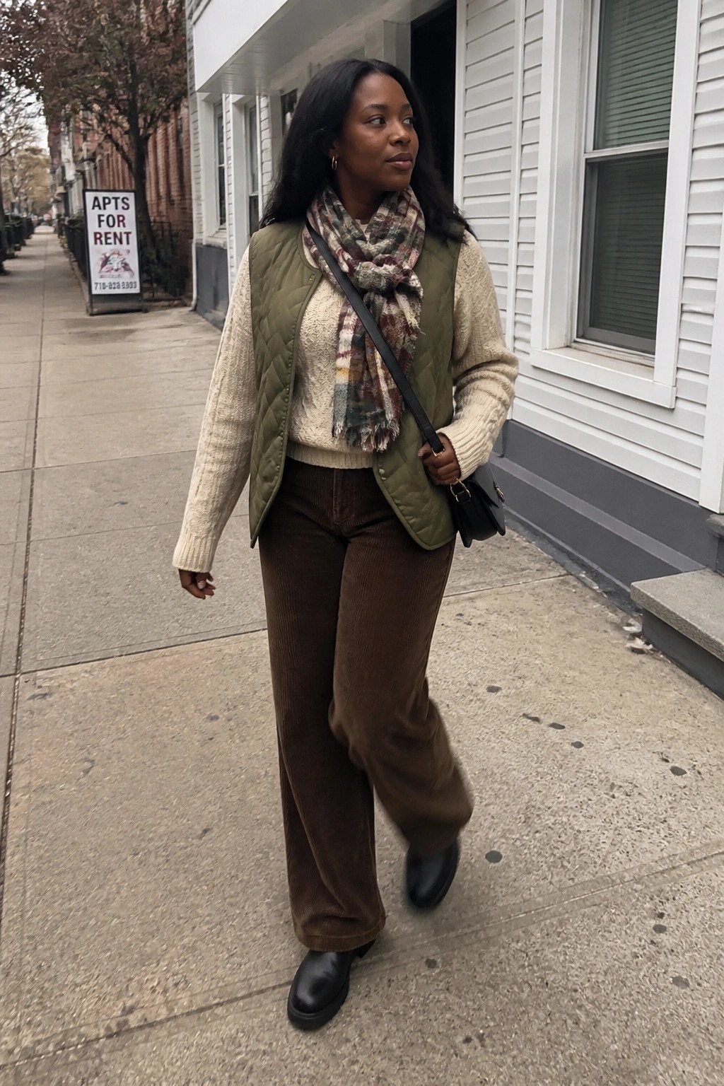 Woman in green quilted vest, plaid scarf, and brown corduroy pants on sidewalk.