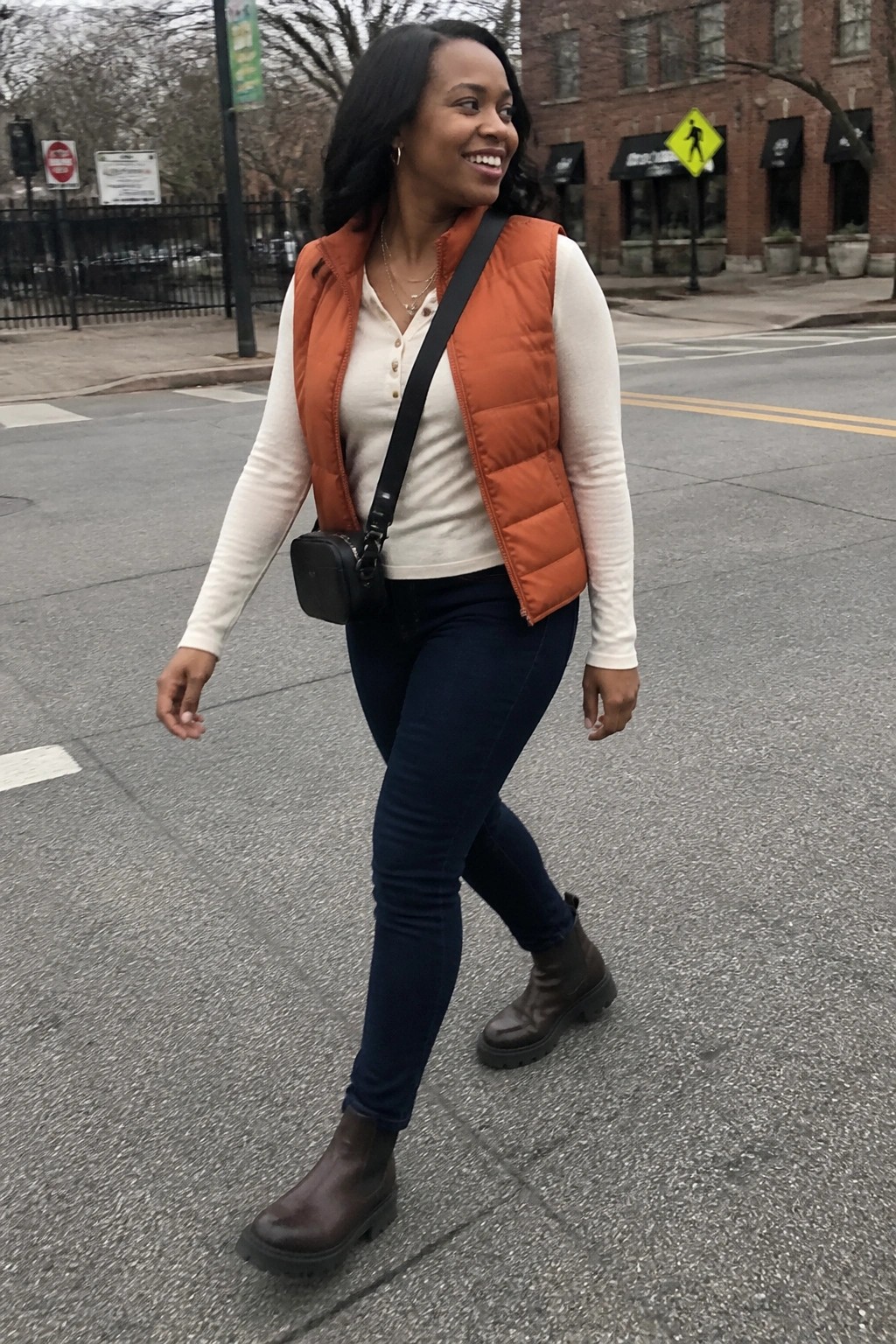 Woman in orange puffer vest and jeans walks on city street smiling