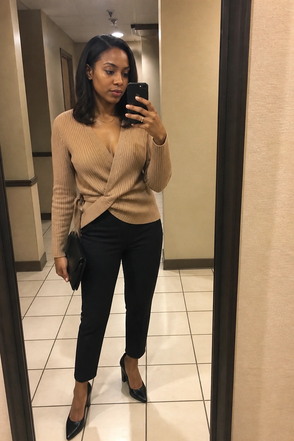 Woman in tan wrap sweater and black pants taking mirror selfie in hallway