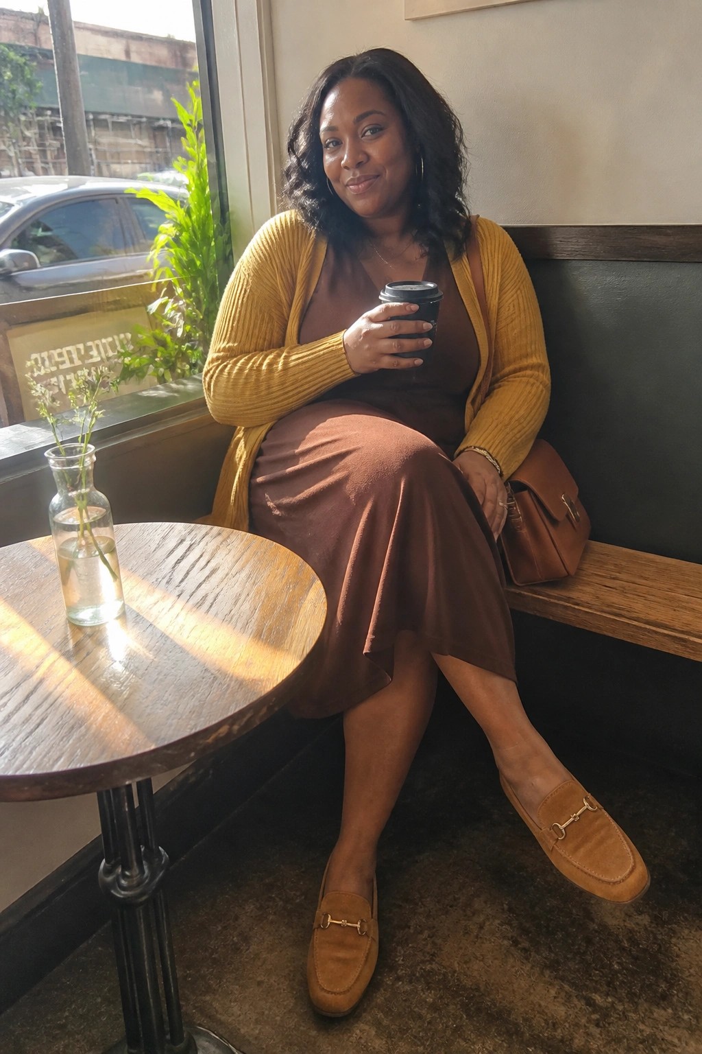Woman in yellow cardigan and brown dress holds coffee in sunlit cafe booth.