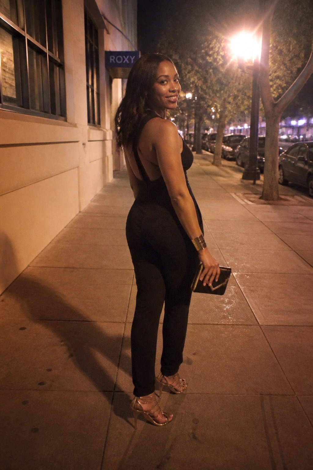 Woman in black backless jumpsuit with gold heels on nighttime sidewalk.