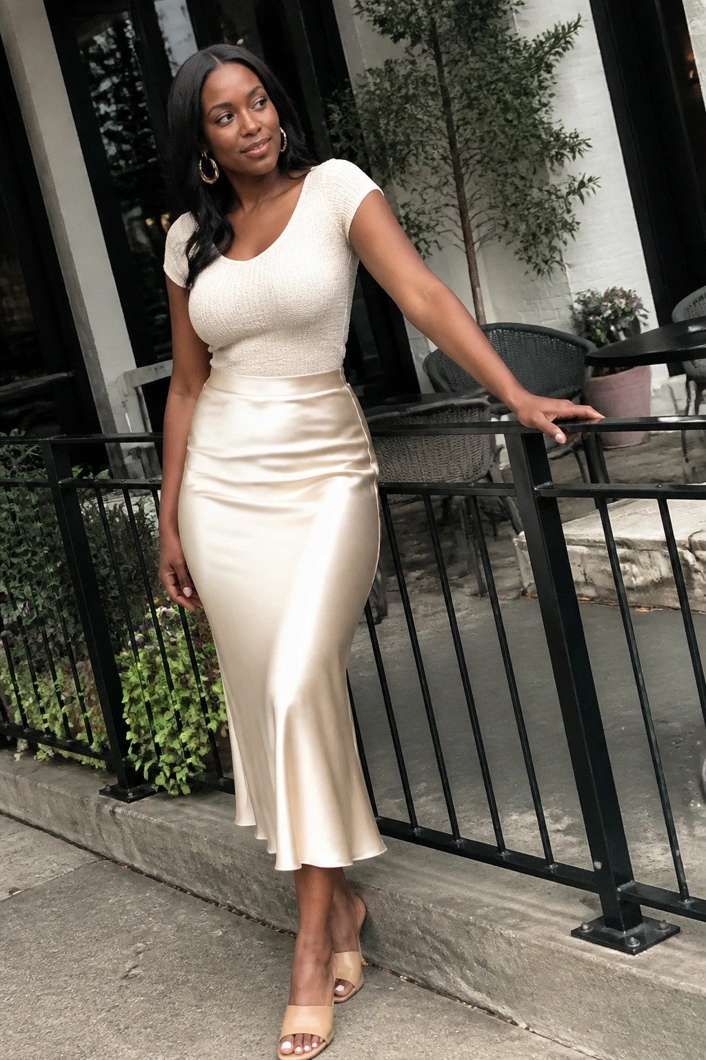 Woman in cream knit top and satin midi skirt leaning on outdoor railing