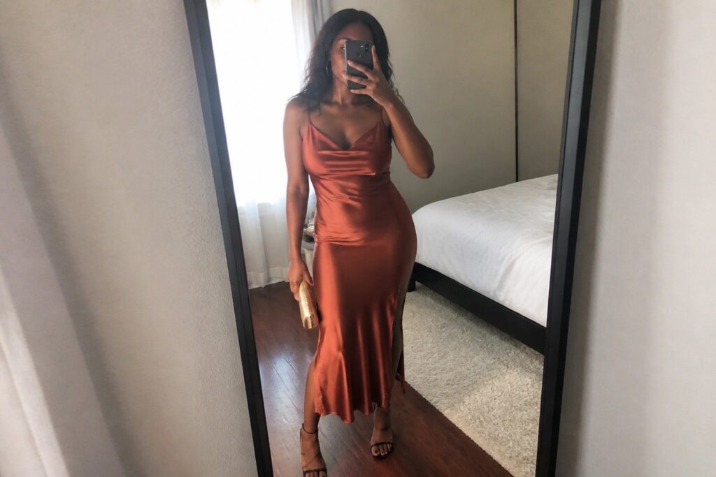 20 Date Night Outfits for Black Women That Feel Chic and Romantic Woman wearing a burnt orange satin midi slip dress with cowl neck and asymmetrical hem paired with black strappy heels and a gold clutch while taking a mirror selfie in a bedroom.