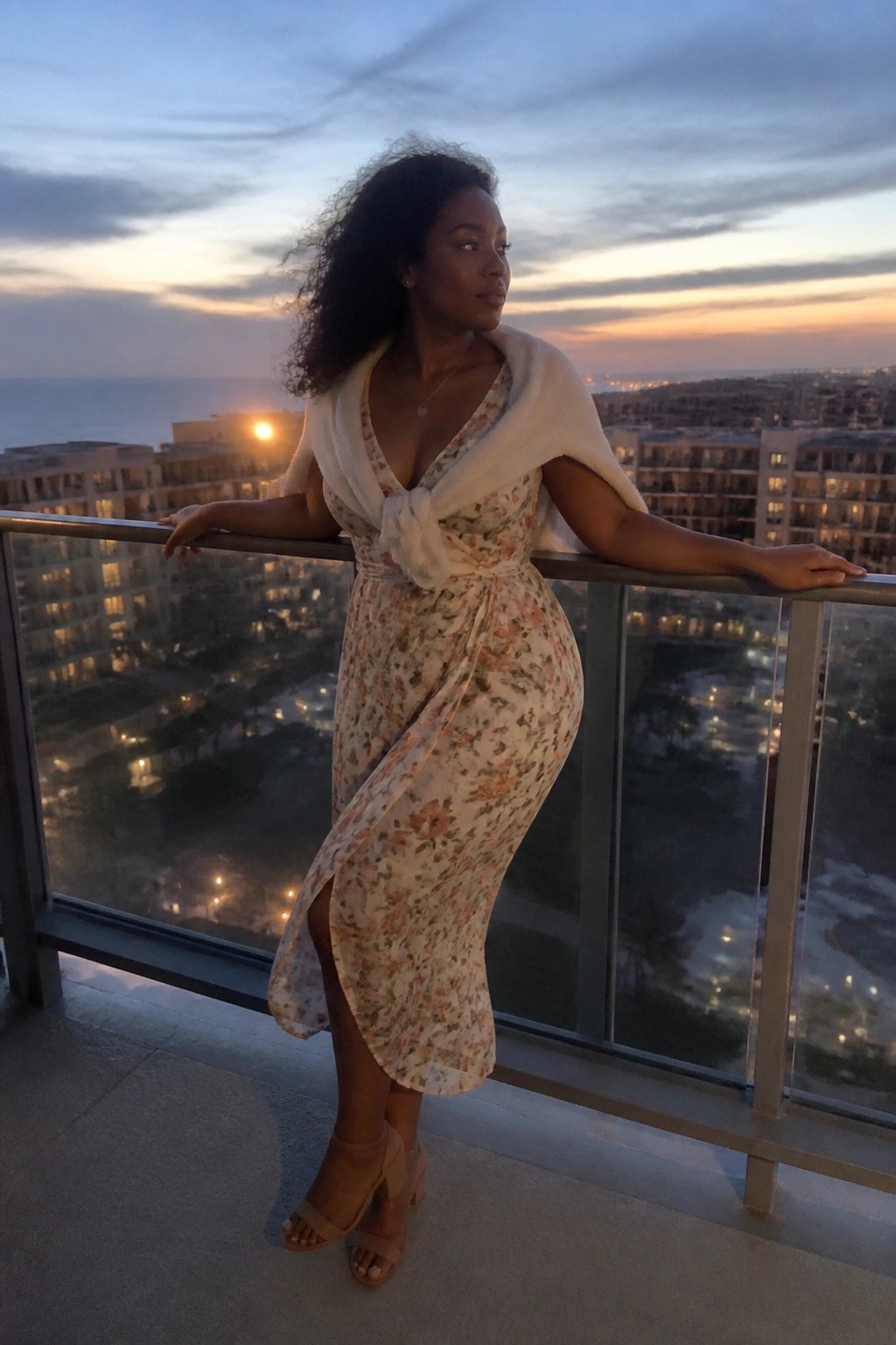 Woman in floral dress and white shawl leaning on balcony at sunset