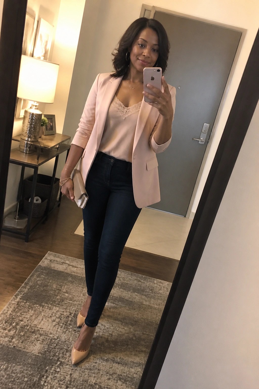 Woman wearing a light pink blazer over a pink lace camisole with dark skinny jeans and nude pointed toe heels while holding a phone and clutch in a mirror selfie.