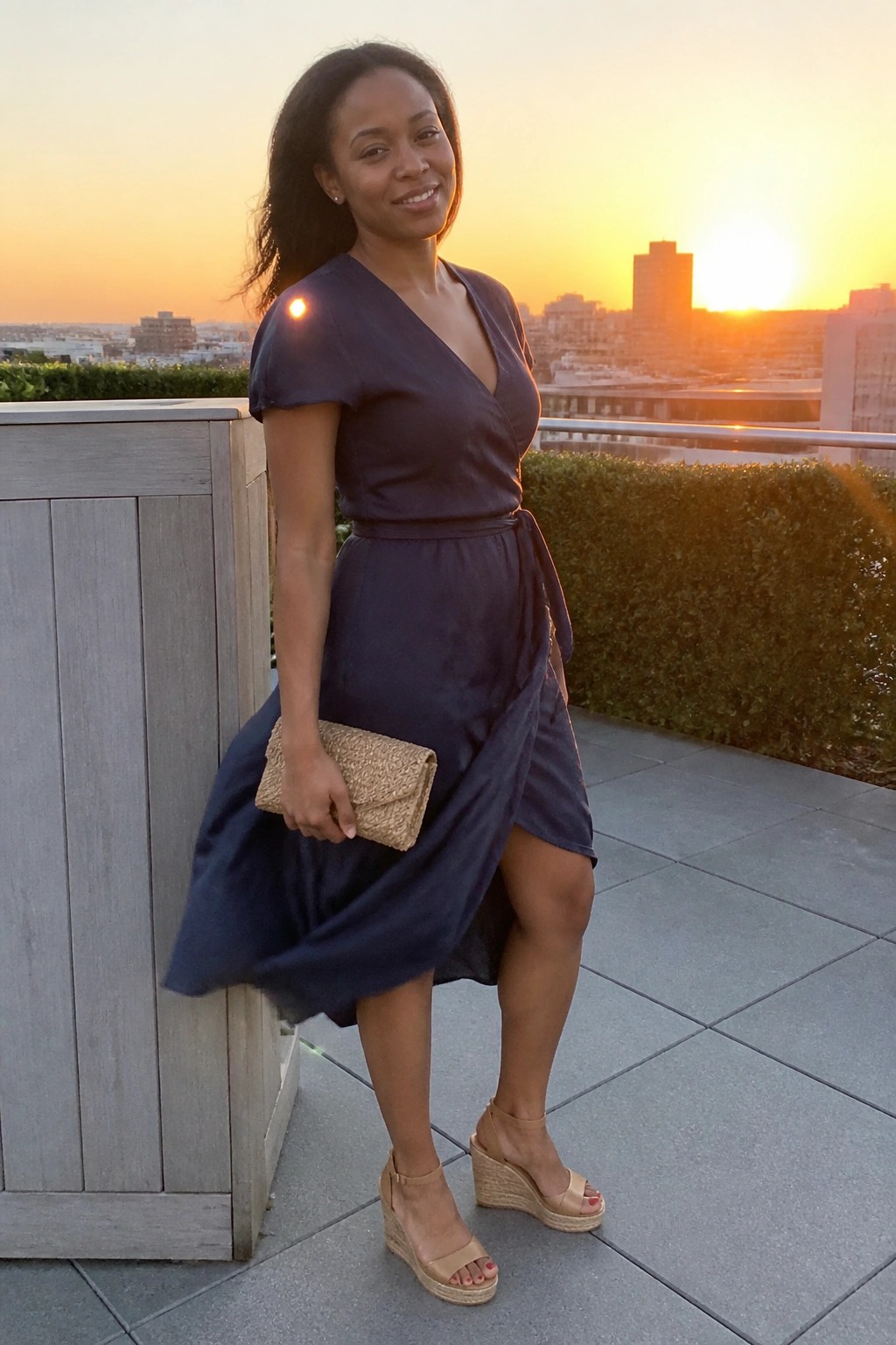 Woman in navy wrap dress holding clutch on rooftop at sunset