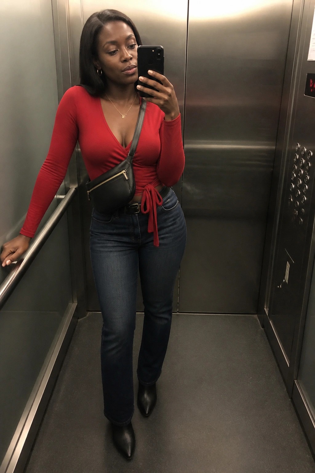 A woman in a red wrap top tied at the waist with straight leg blue jeans black pointed boots and a black crossbody bag taking a mirror selfie inside an elevator.