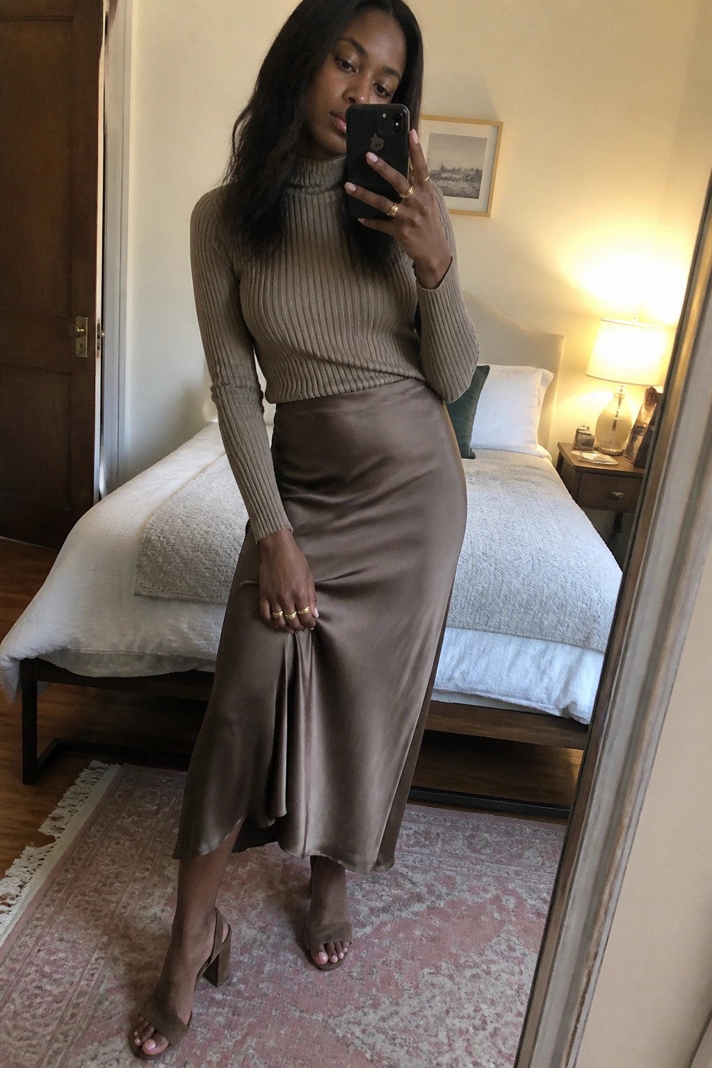 Woman in taupe ribbed turtleneck and satin midi skirt taking bedroom mirror selfie.