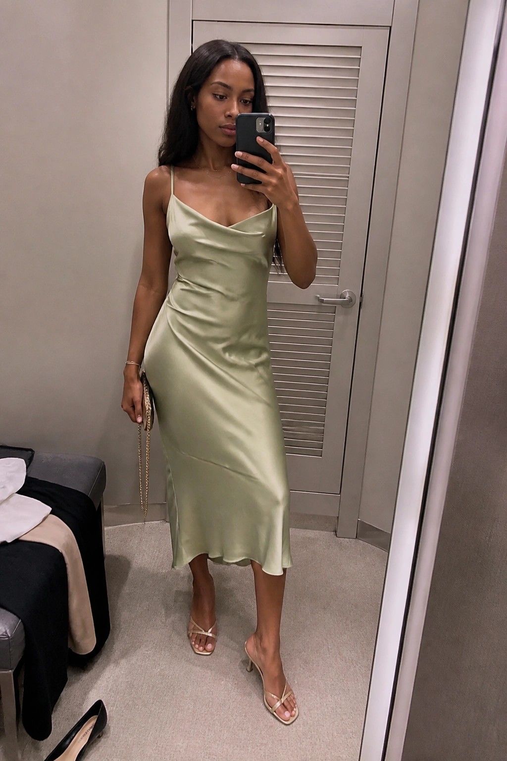 A woman in a sage green satin midi slip dress with thin straps and cowl neckline, gold strappy heels, and a gold chain purse, shown in a full-length mirror selfie.