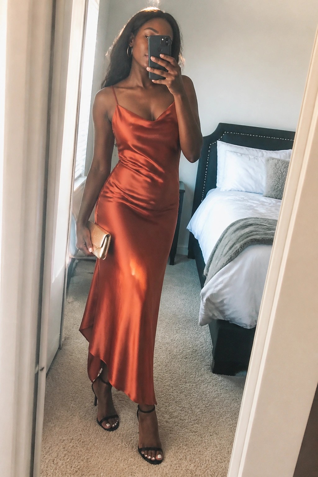 Woman wearing a burnt orange satin midi slip dress with cowl neck and asymmetrical hem paired with black strappy heels and a gold clutch while taking a mirror selfie in a bedroom.