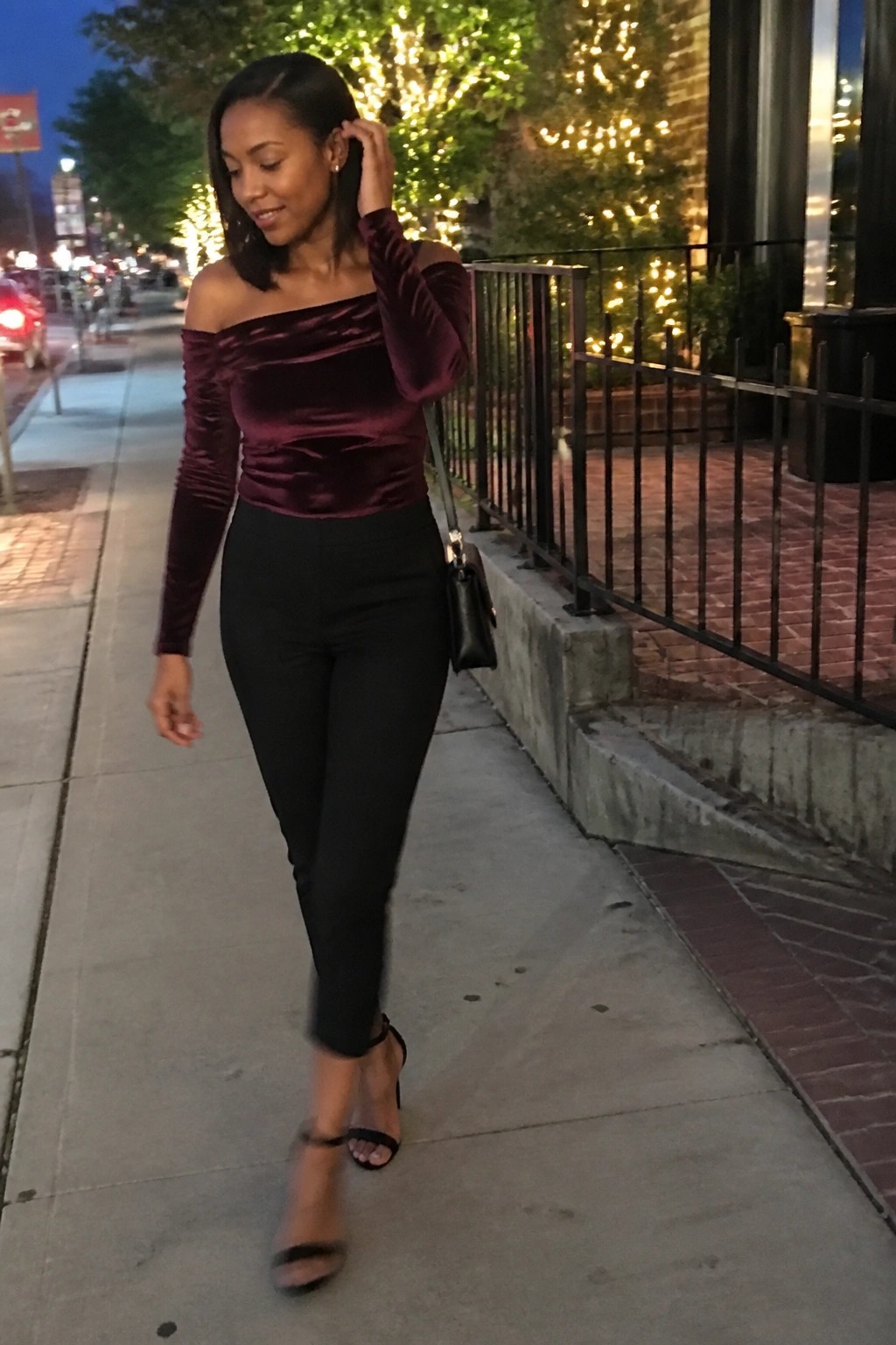 Woman in burgundy velvet off shoulder long sleeve top black slim pants and black strappy heels carrying small black handbag on city sidewalk.