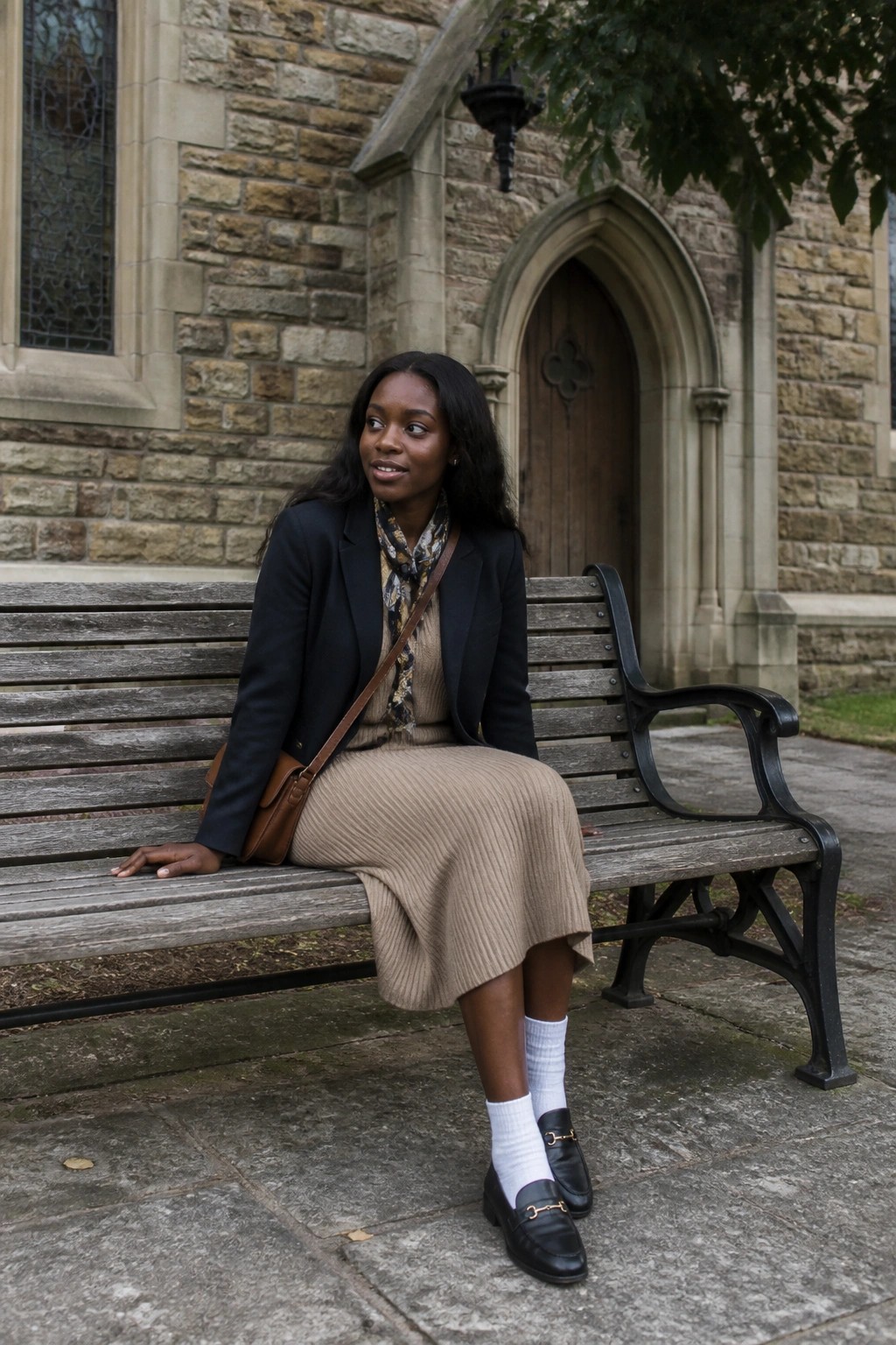 Woman in black blazer and pleated skirt on bench outside stone building.