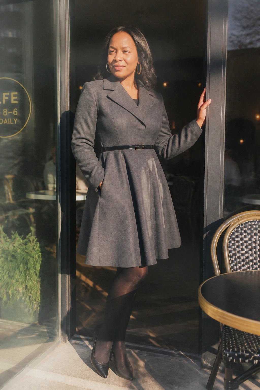 Woman wearing a gray belted A-line coat dress with black tights and pointed heels standing in a doorway.