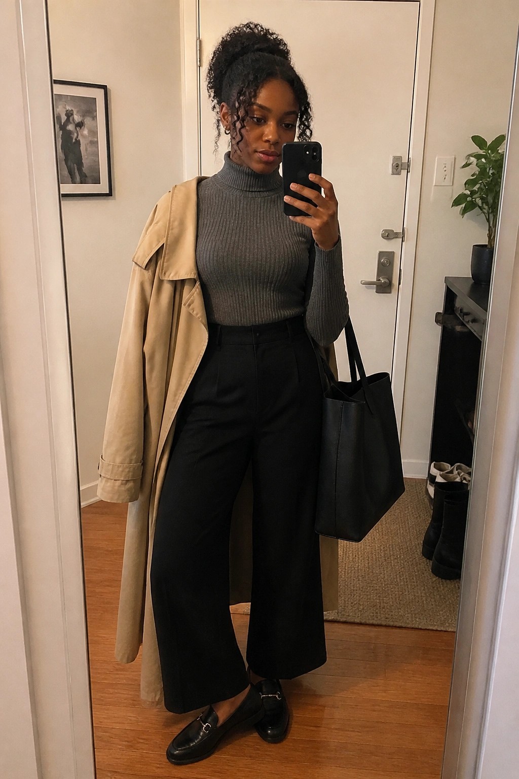 Woman in gray turtleneck, black pants, and beige trench coat holding phone and tote bag