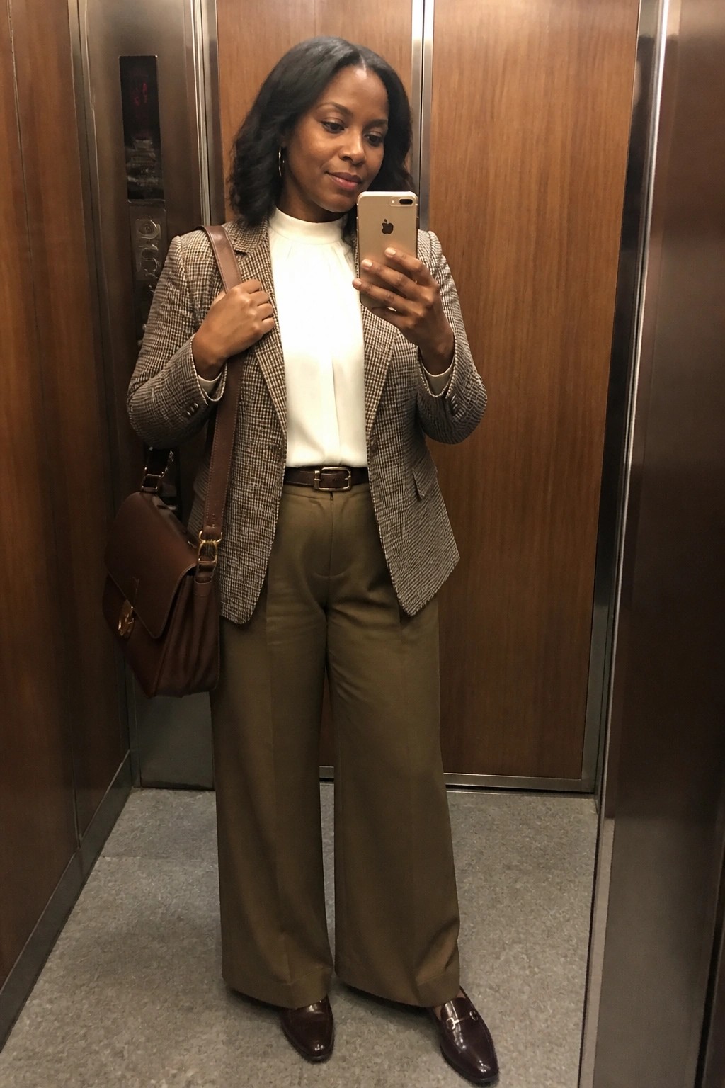 A Black woman stands in an elevator taking a mirror selfie while wearing a brown and white houndstooth blazer over a white blouse paired with olive green wide leg pants a brown belt brown loafers and a brown shoulder bag.