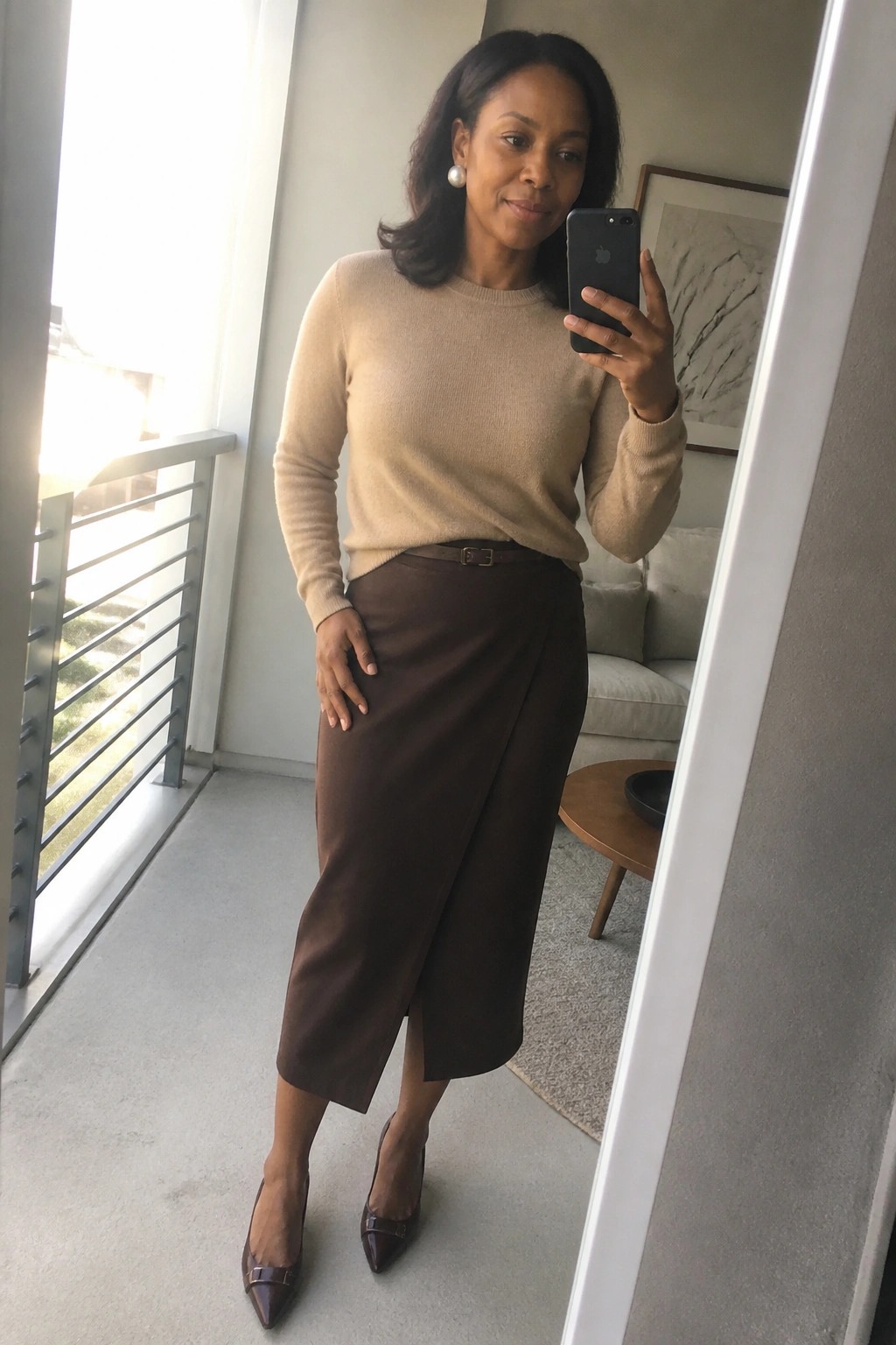 A Black woman wearing a beige knit sweater tucked into a brown midi wrap skirt with a matching belt, brown pointed heels, and pearl earrings while taking a mirror selfie.