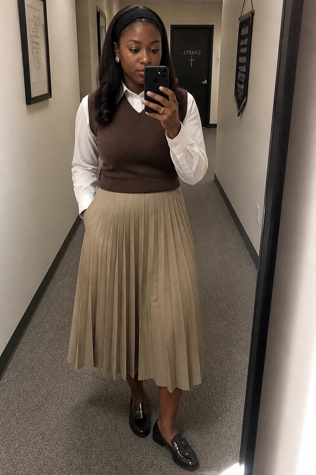 Woman in brown sweater vest and beige pleated skirt taking mirror selfie in hallway