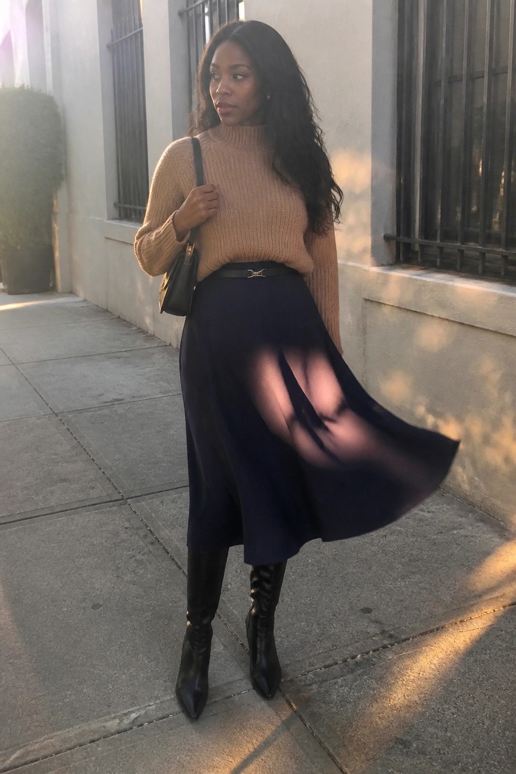 Woman in beige turtleneck sweater and flowing navy skirt with black boots on sidewalk.