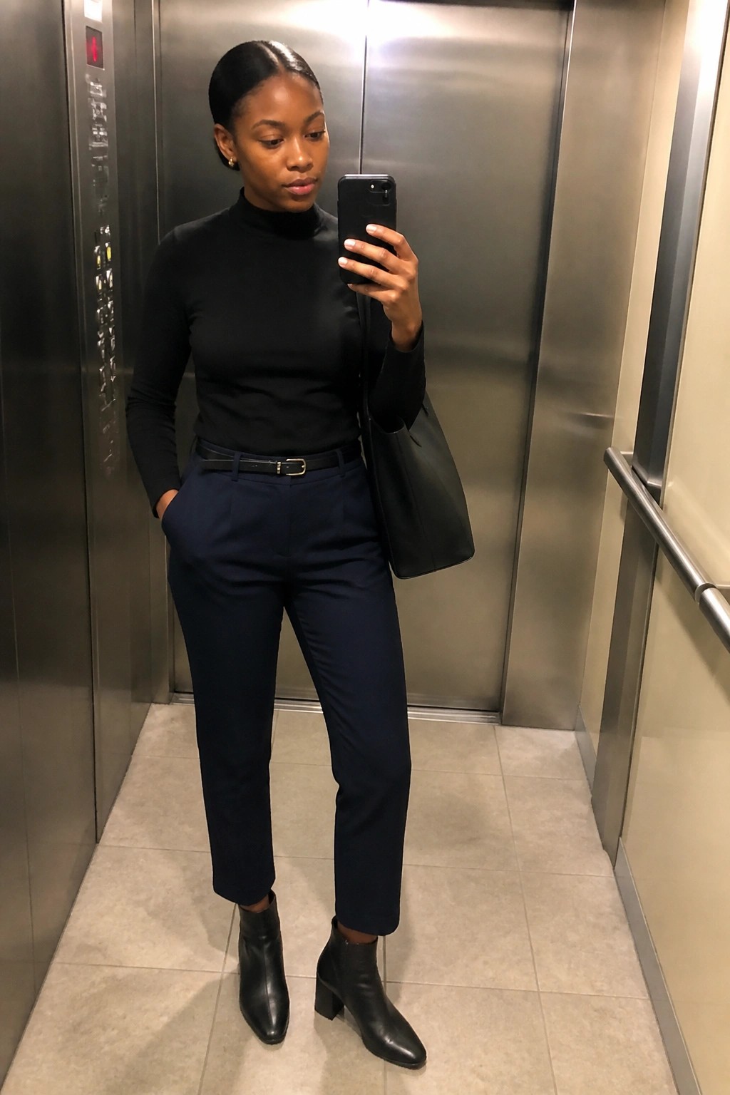 Woman in black turtleneck and navy pants taking selfie in elevator