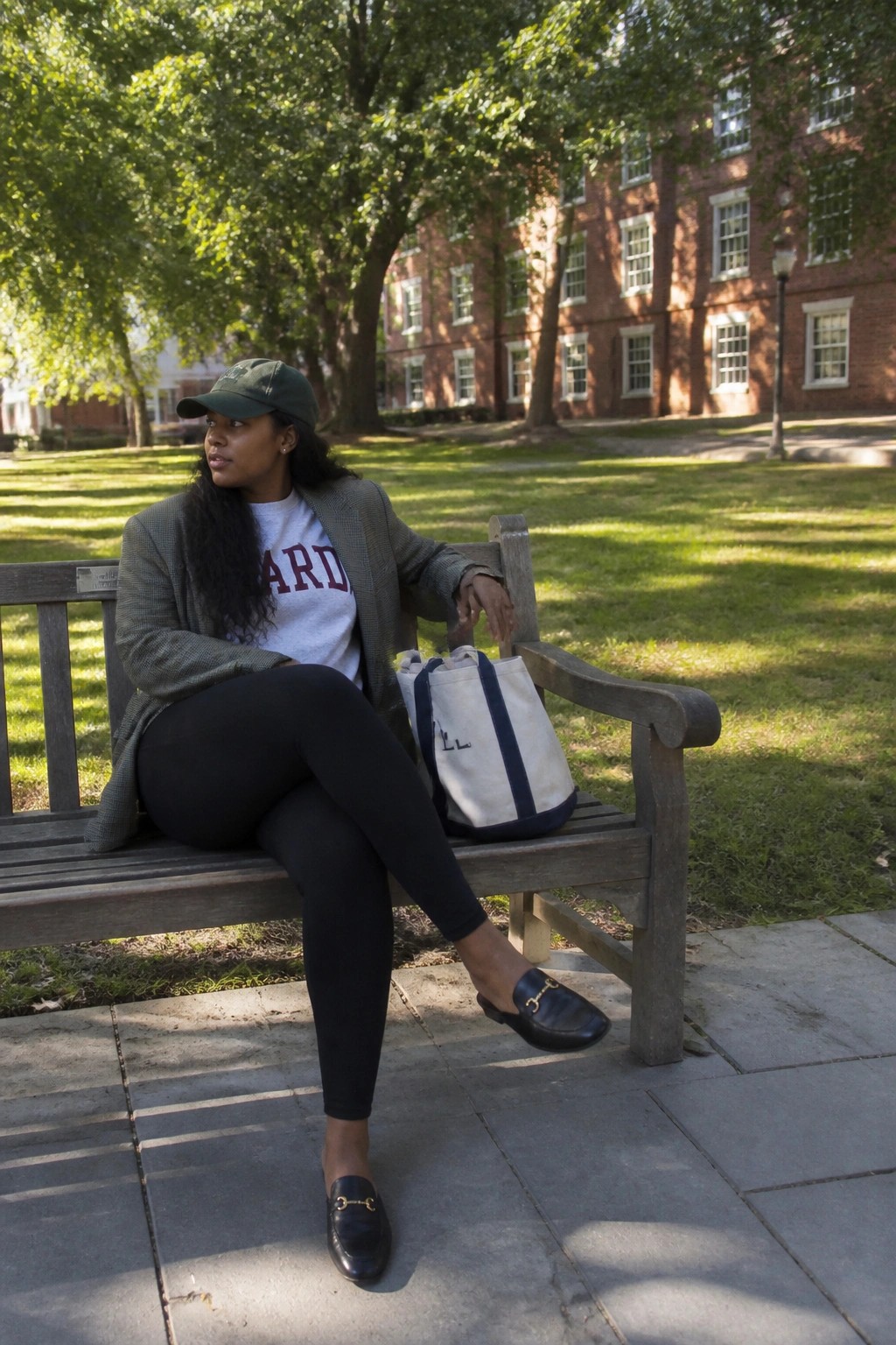 Woman in Harvard shirt and blazer seated on campus bench with tote bag