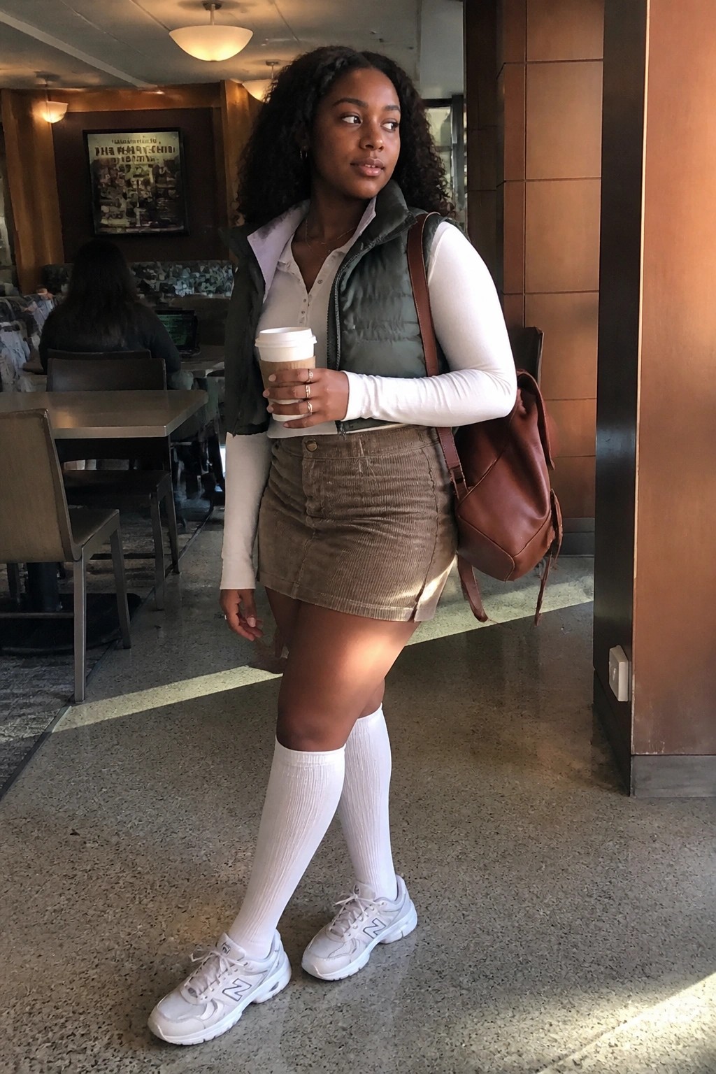 Woman in white long sleeve button down shirt under green puffy vest, brown corduroy mini skirt, white knee high socks, white sneakers, and brown leather backpack holding a coffee cup.