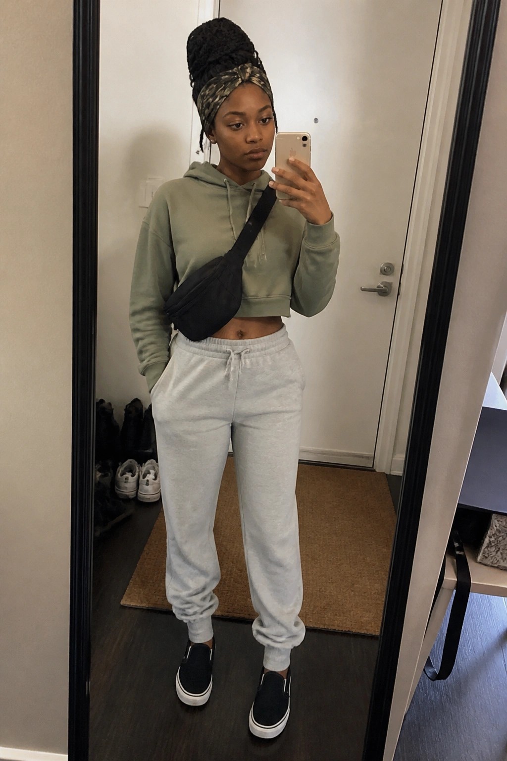 Woman in sage green cropped hoodie and gray sweatpants taking mirror selfie with crossbody bag