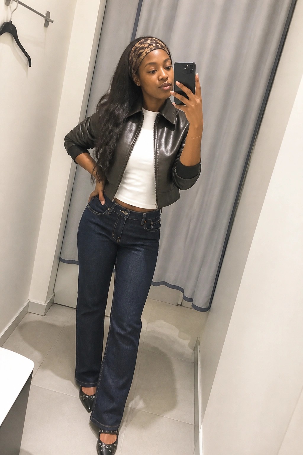 Woman in black leather jacket and blue jeans taking mirror selfie in fitting room.