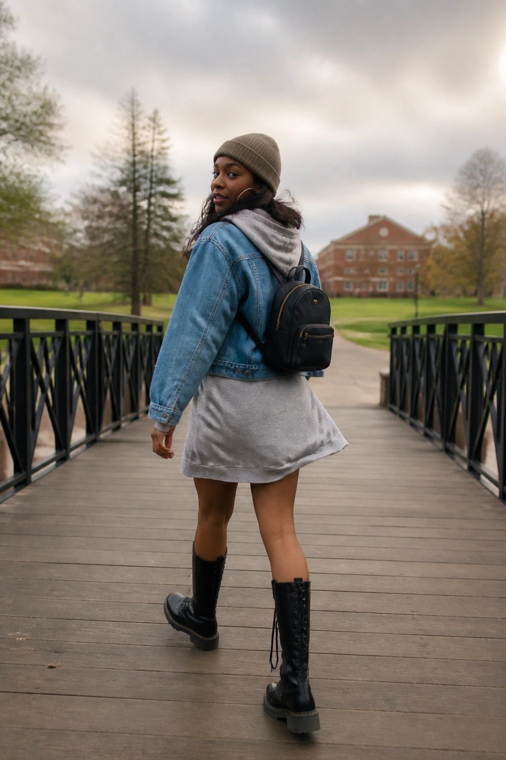 A Black woman in a blue denim jacket layered over a grey hoodie and grey sweatshirt dress, black combat boots, black mini backpack, and beige beanie.