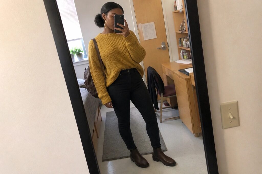 Woman in yellow sweater, black jeans, and brown boots taking mirror selfie in bedroom.