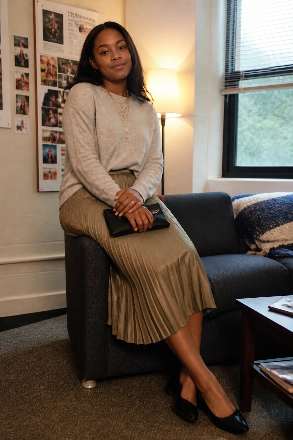Woman in beige sweater and pleated skirt sits on couch holding black clutch.