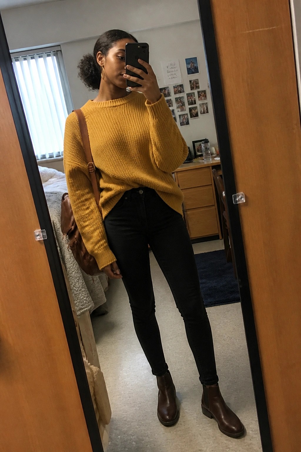 Woman in yellow sweater, black jeans, and brown boots taking mirror selfie in bedroom.