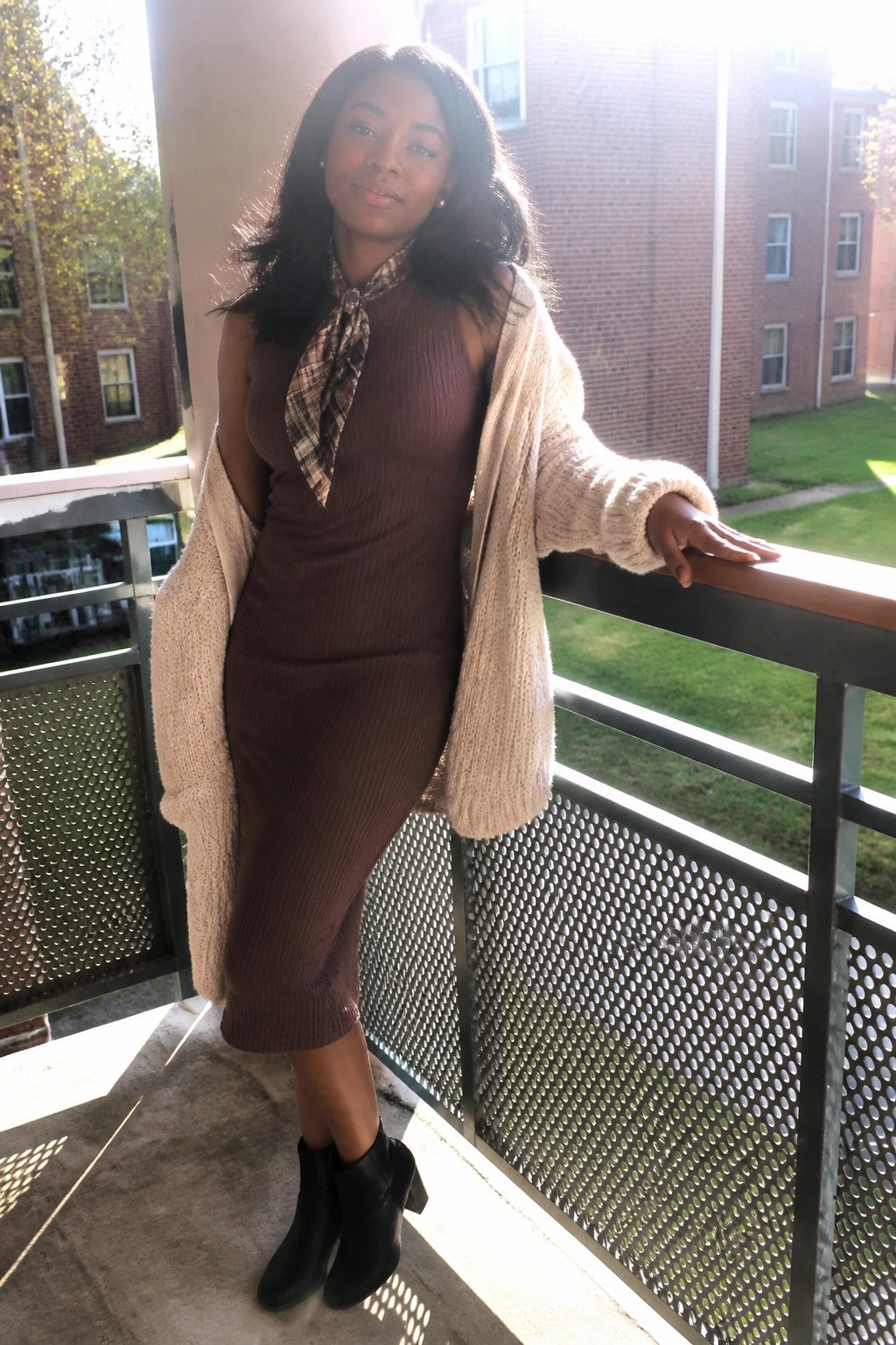 Woman in brown ribbed dress with plaid scarf and beige cardigan on sunny balcony