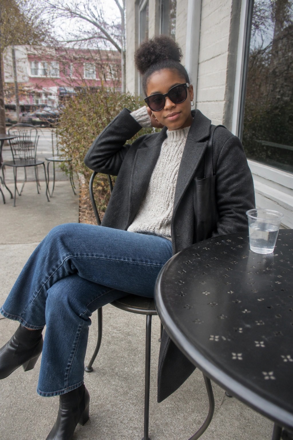 A Black woman seated outdoors wearing blue wide-leg jeans, a cream chunky knit sweater, an open dark gray coat, black ankle boots, and dark sunglasses with her hair in a high bun.