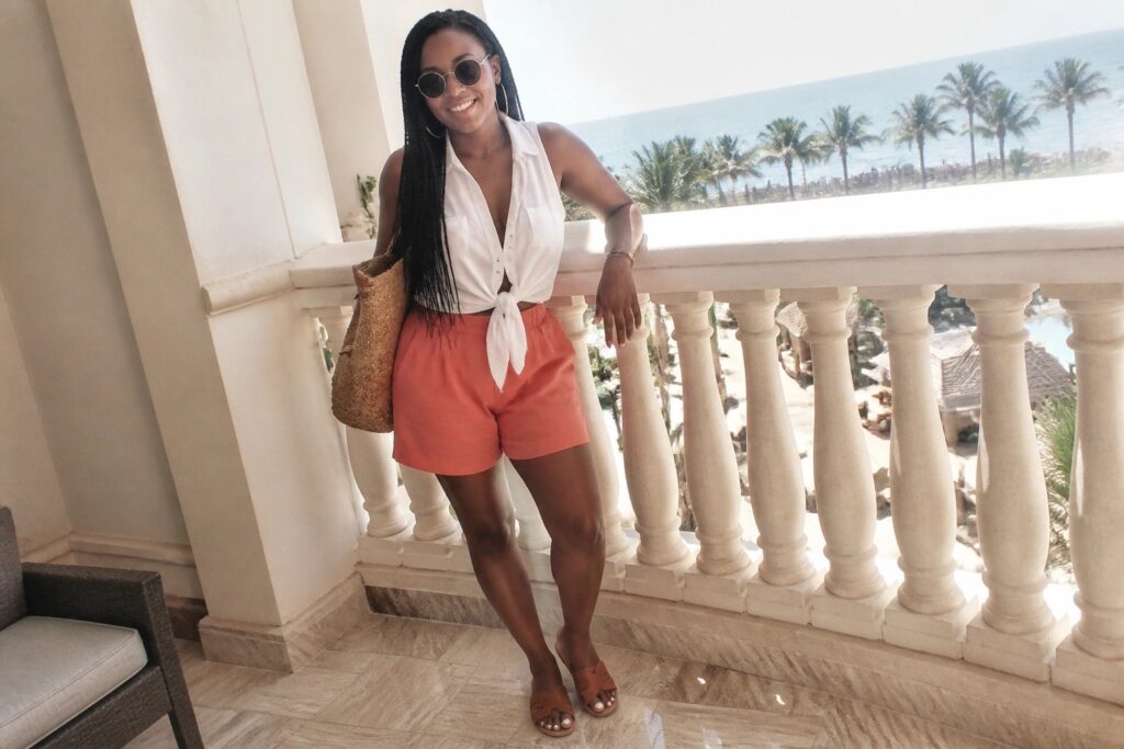 Woman in white knotted shirt and coral shorts on ocean-view balcony with bag