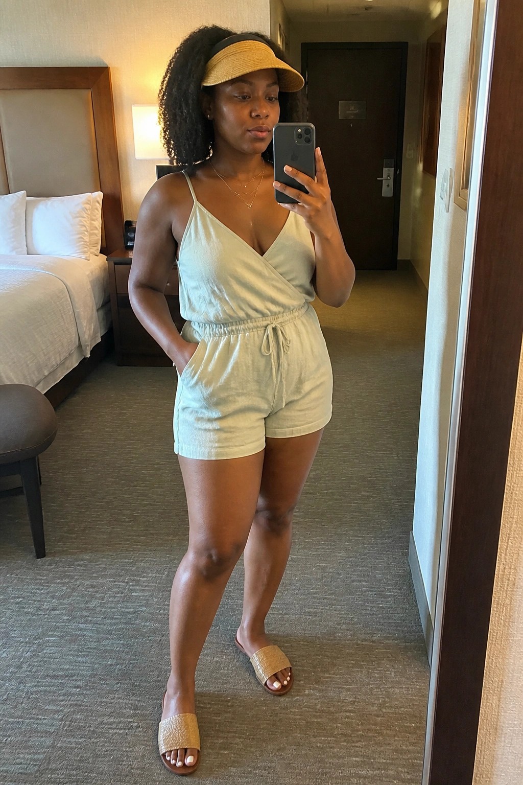 Woman in light green romper and yellow visor taking mirror selfie in hotel room