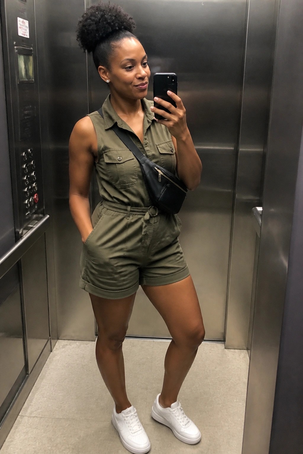 Woman in olive green romper taking mirror selfie in elevator with white sneakers