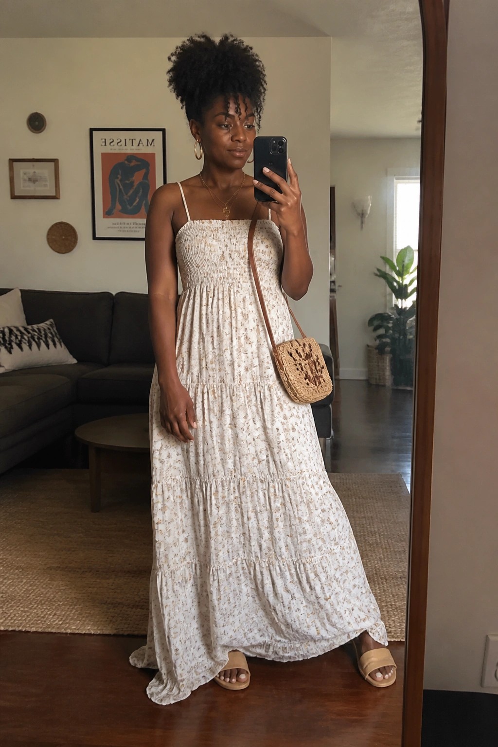 Woman wearing a white tiered maxi dress with small brown floral print, thin straps, smocked bodice, woven straw bag on leather strap, beige slide sandals, and gold jewelry, standing indoors in front of a mirror.