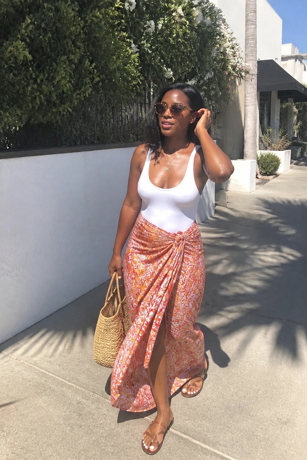 Woman in white bodysuit and orange patterned wrap skirt holding woven bag outdoors
