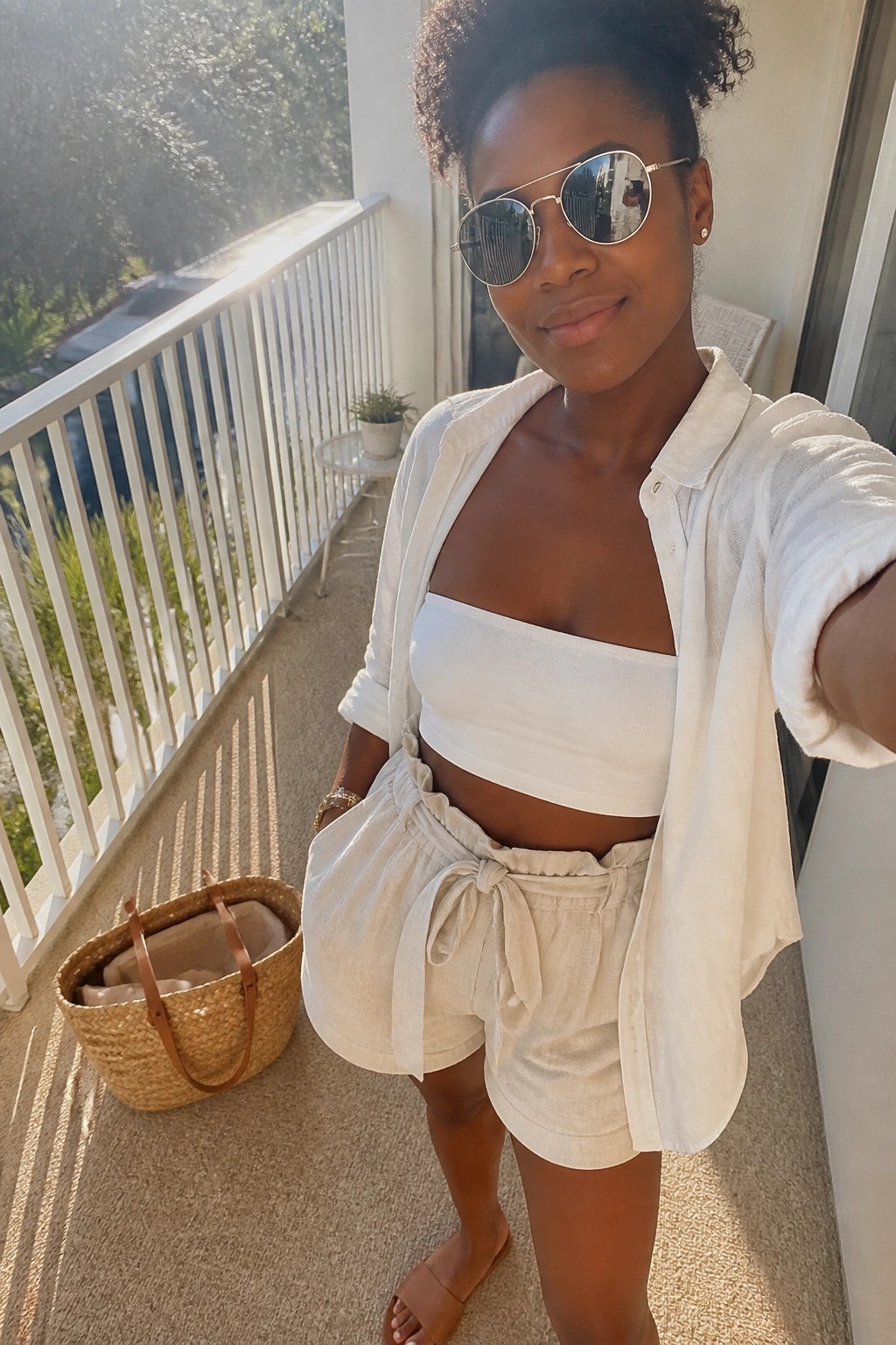 A woman wearing an open white linen button-up shirt over a white bandeau crop top and matching white linen shorts with a front tie, accessorized with brown slide sandals, a woven straw tote, and aviator sunglasses.